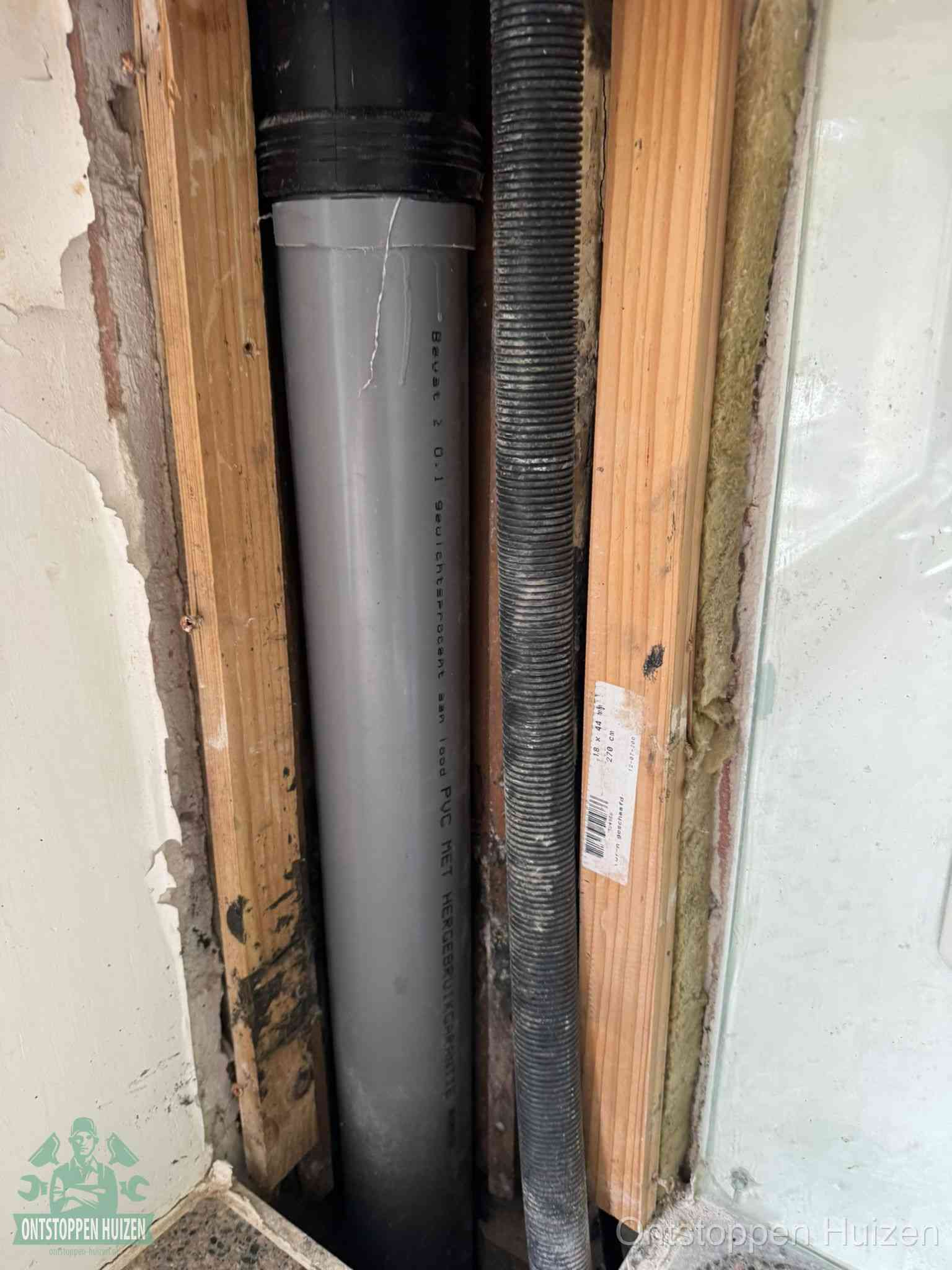 Een grijze PVC-afvoerbuis met een flexibele ribbelslang in een open wandconstructie, een verwaarloosde afvoer die te lang is blijven zitten in Huizen