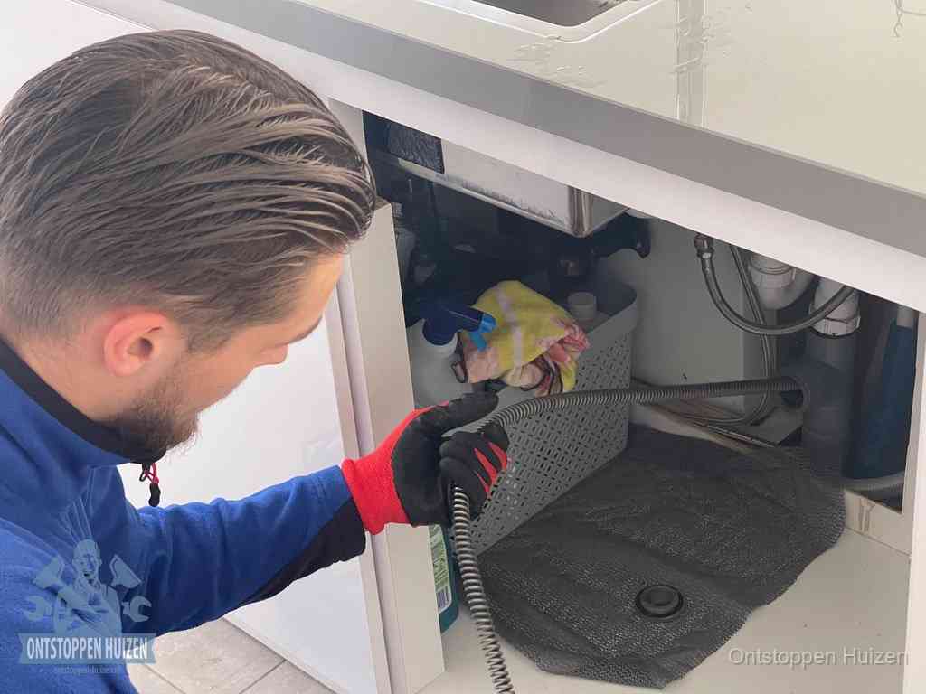 Een monteur voert een flexibele ontstoppingsveer in onder het keukenkastje, langs de sifon, om de gootsteen te ontstoppen in Huizen
