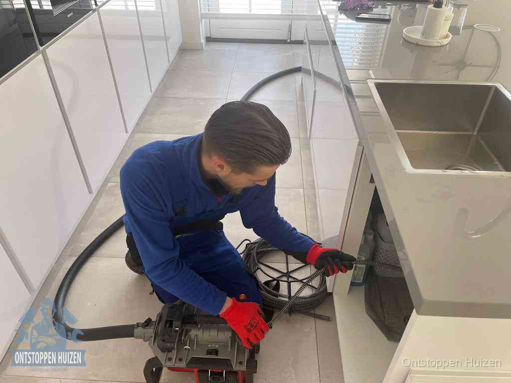 Een monteur ontstopt de keukenafvoer onder de spoelbak met een elektrische veermachine in Huizen