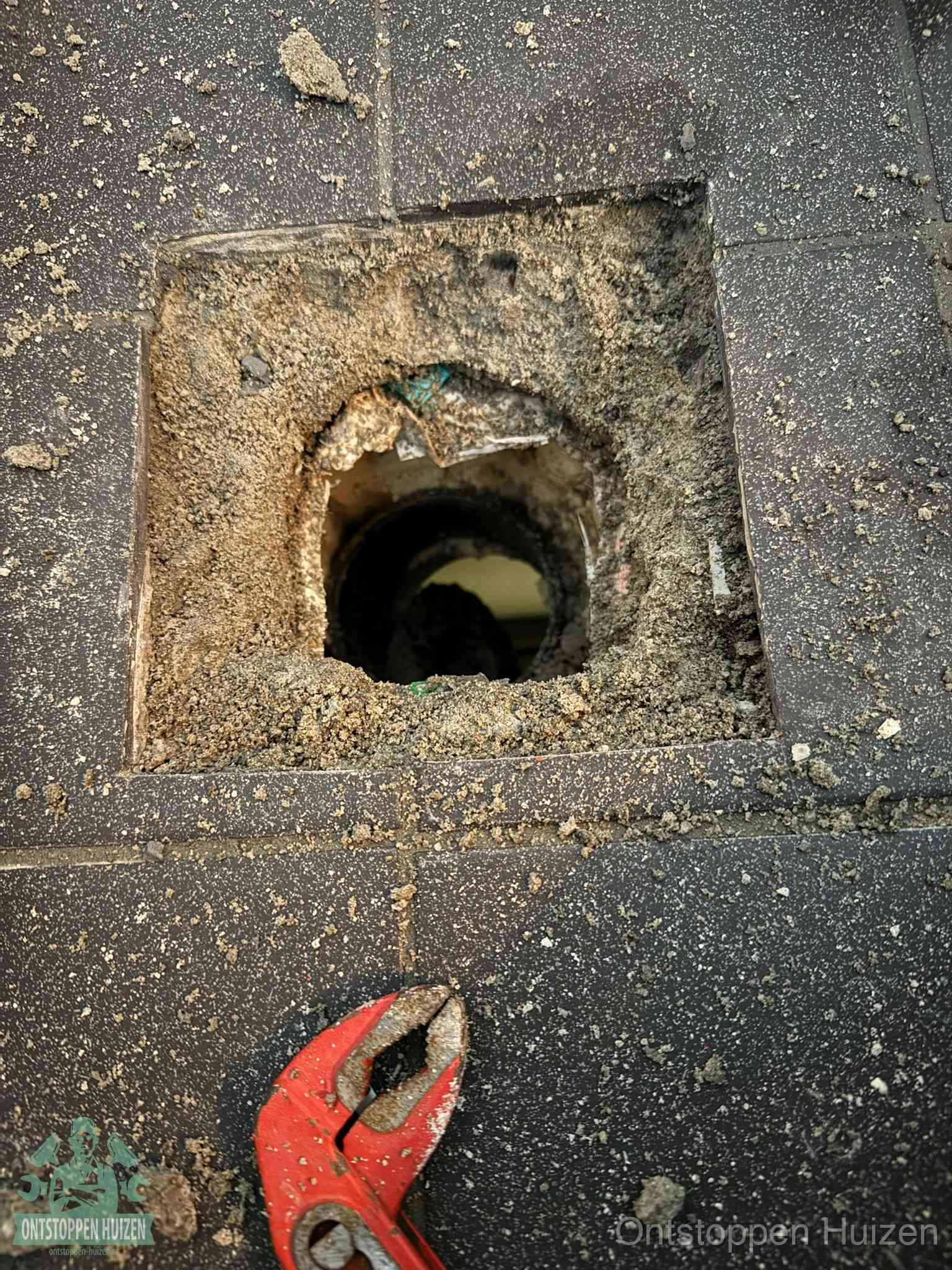 Een geopend rioolputje in een betontegelvloer met losgemaakt rooster en een zichtbare ronde afvoerbuis, het achterhalen waar de afvoerverstopping zit in Huizen