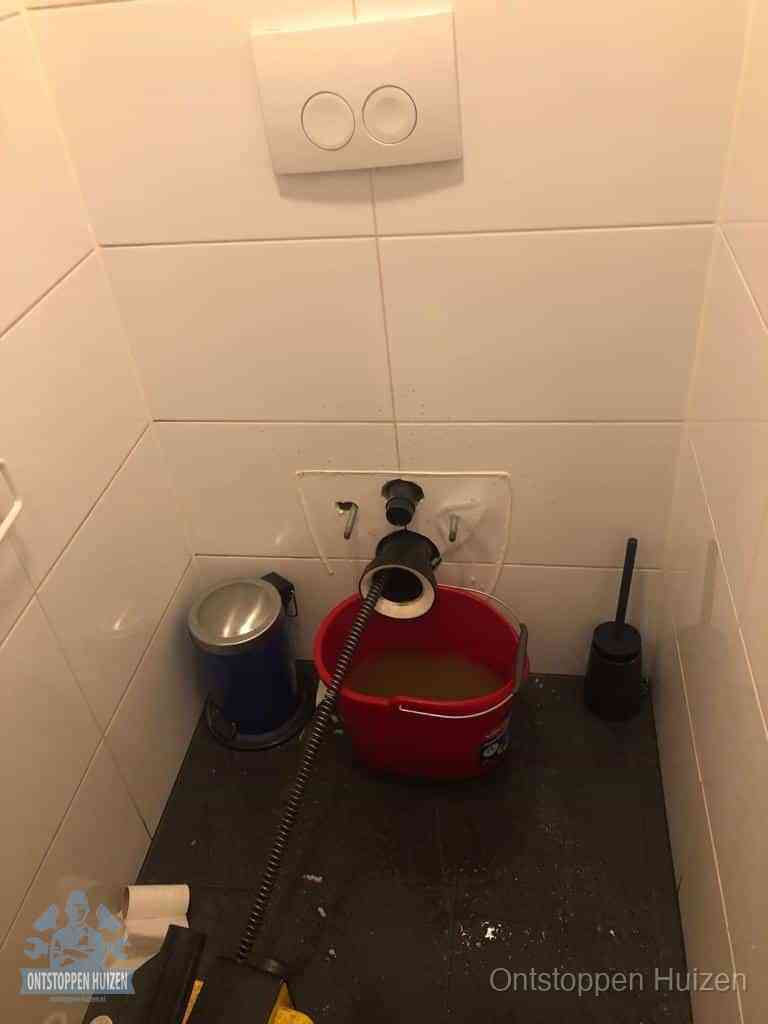 Een ontstopte wand-wc met verwijderde toiletpot waar een ontstoppingsveer in de wanduitgang verdwijnt, het achterhalen waar de verstopping zit in Huizen