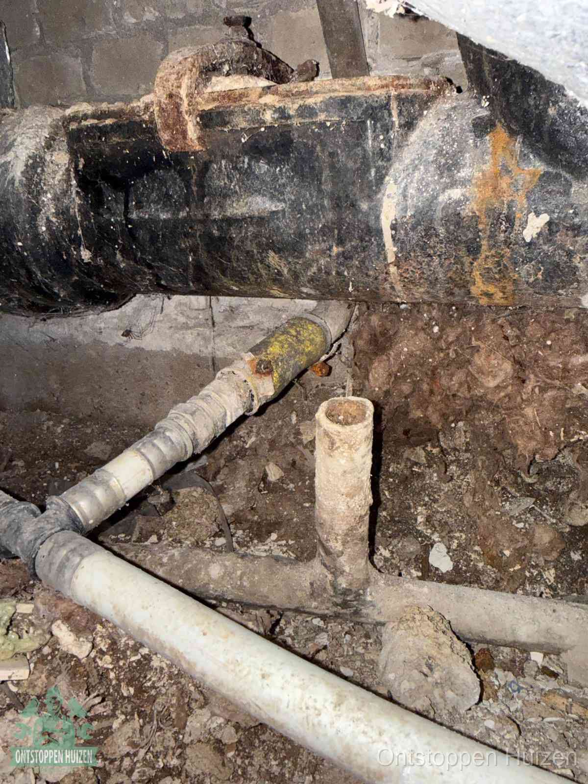 Een dikke zwarte hoofdrioolbuis met roest- en kalkaanslag in een kruipruimte, oorzaak van terugkerende structurele afvoerproblemen in Huizen