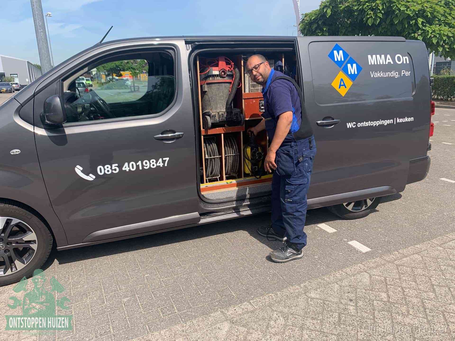 Een monteur bij de open schuifdeur van zijn bedrijfsbus op een parkeerplaats, klaar om de oorzaak van terugkerende afvoerverstoppingen aan te pakken in Huizen