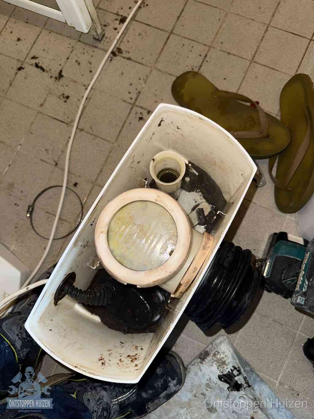 Een gedemonteerde toiletpot op de vloer met ontstoppingsveer en gereedschap ernaast, het verwijderen van een vastzittend voorwerp uit de wc in Huizen