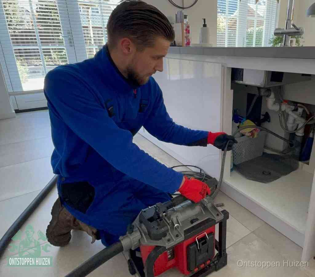 Een monteur met rode handschoenen knielt in een keuken en voert een elektrische ontstoppingsveer in tijdens het professioneel afvoer ontstoppen in Huizen