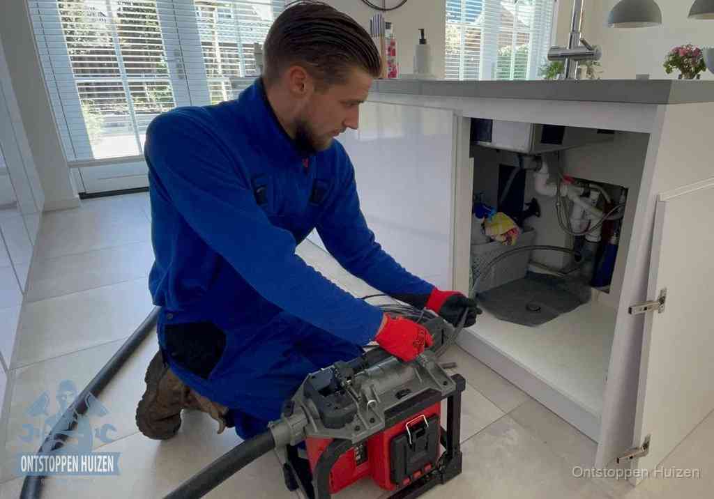 Een monteur voert een professionele elektrische ontstoppingsmachine in de afvoer onder de spoelbak tijdens het gootsteen ontstoppen in Huizen